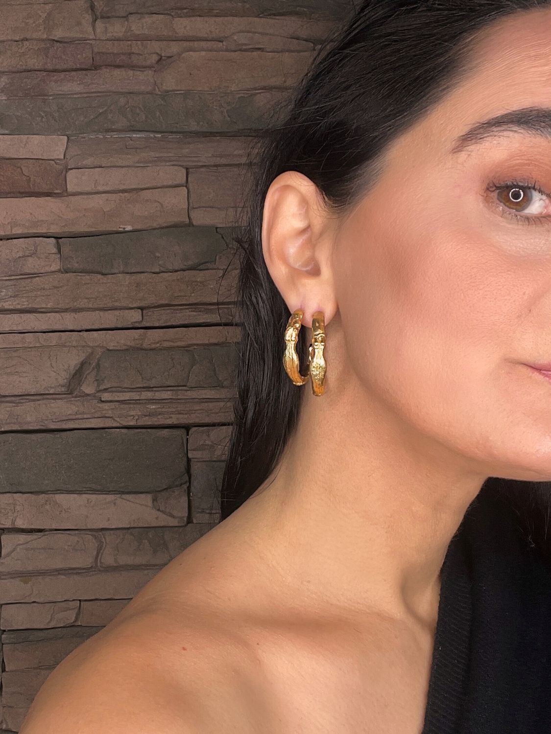 Close-up of a woman wearing goddess gold hoop earrings with a stone wall background