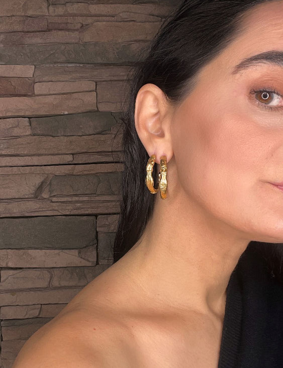 Close-up of a woman wearing goddess gold hoop earrings with a stone wall background