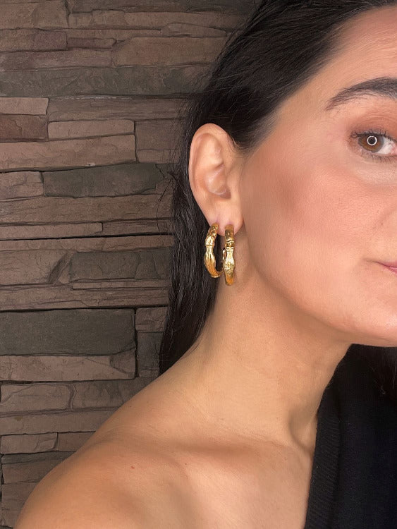 Close-up of a woman wearing goddess gold hoop earrings with a stone wall background