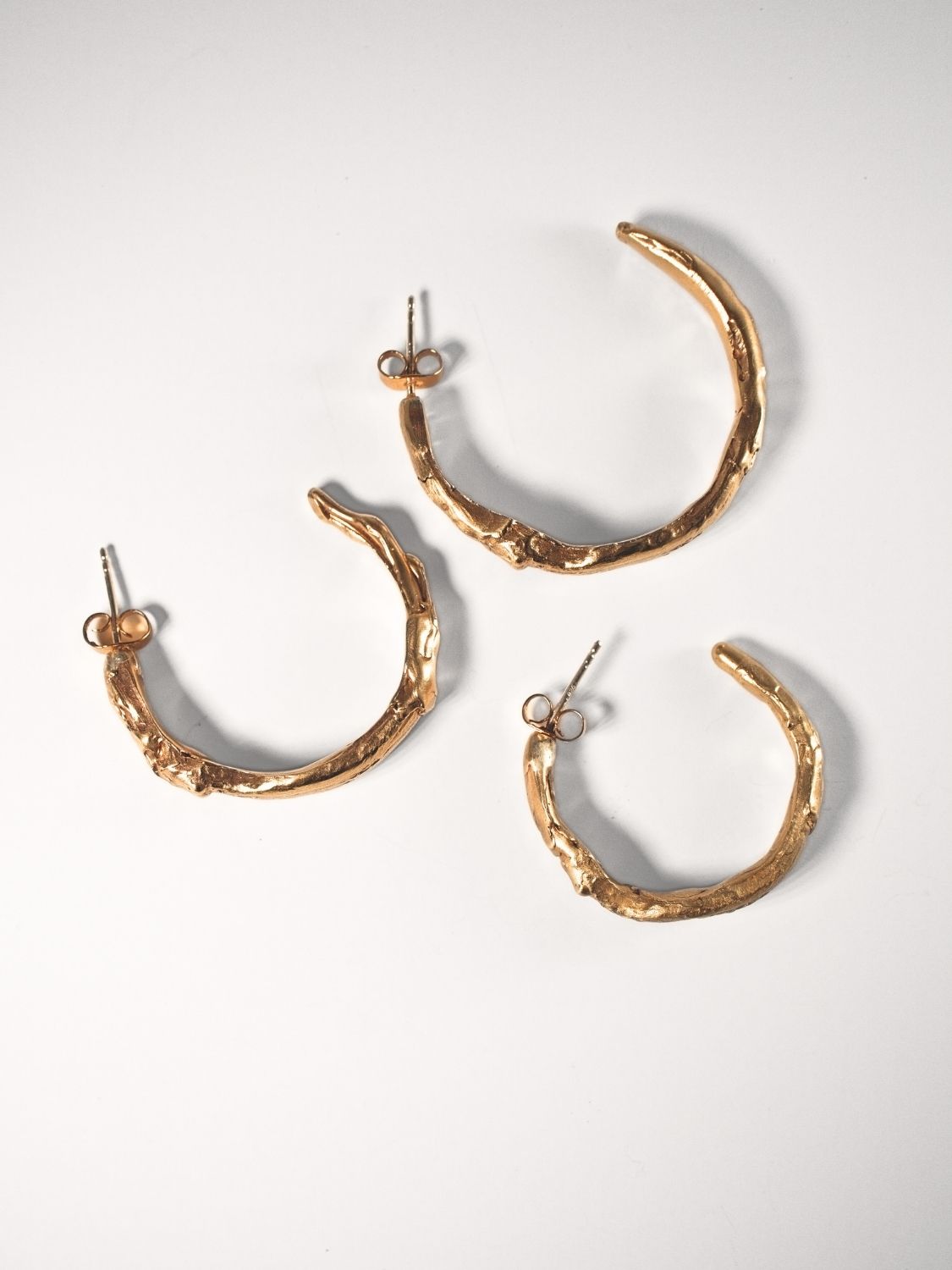 Three sculptural goddess gold hoop earrings on a light grey background