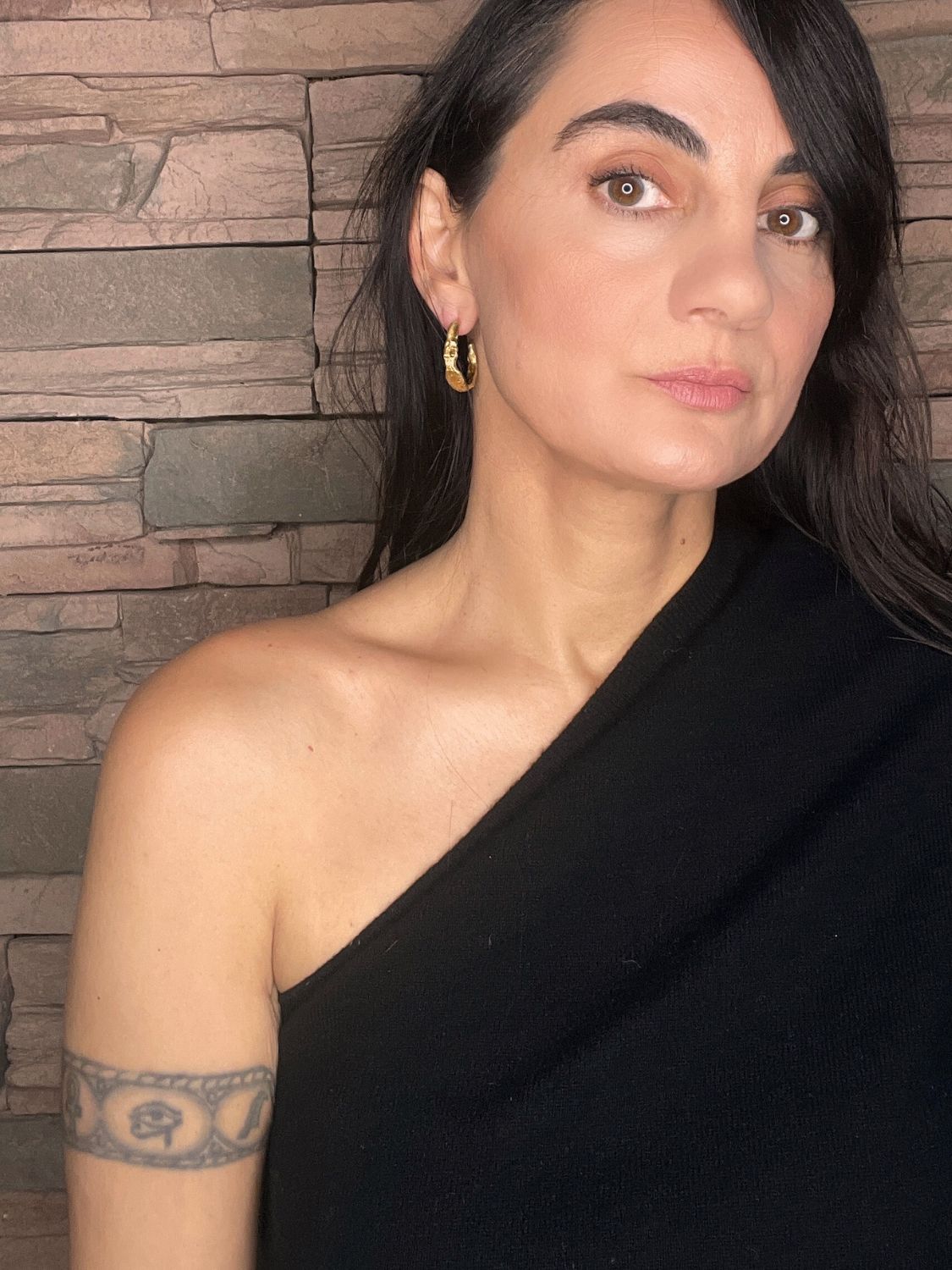 Woman wearing a black top with sculptural gold hoops against a stone wall background