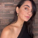 Woman with long dark hair wearing gold goddess hoops and black top against a stone wall background