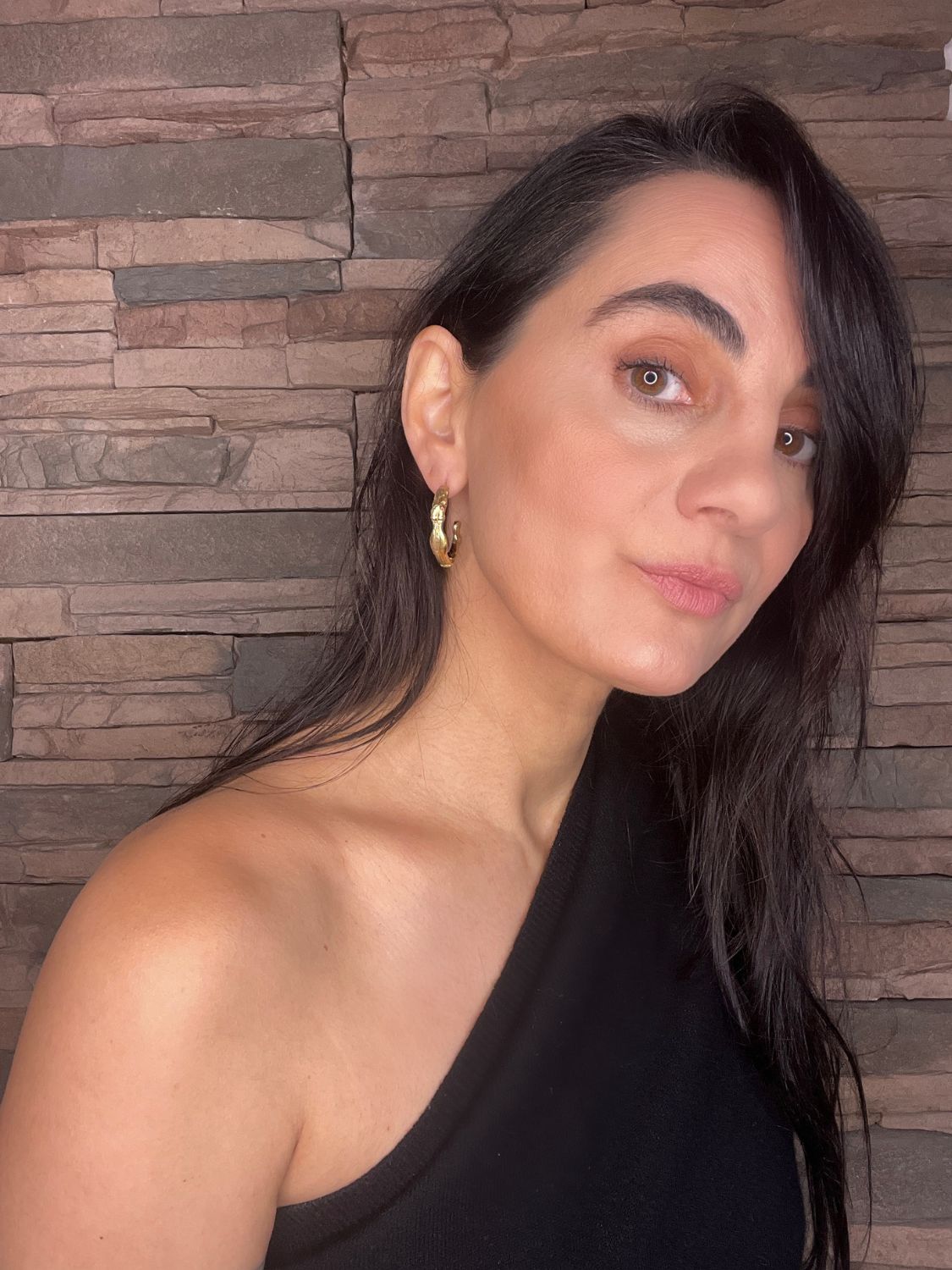 Woman with long dark hair wearing gold goddess hoops and black top against a stone wall background
