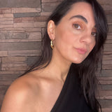 Woman with long dark hair wearing gold goddess hoops and black top against a stone wall background