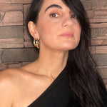 Woman with long dark hair wearing gold sculptural goddess hoop earrings against a stone wall background