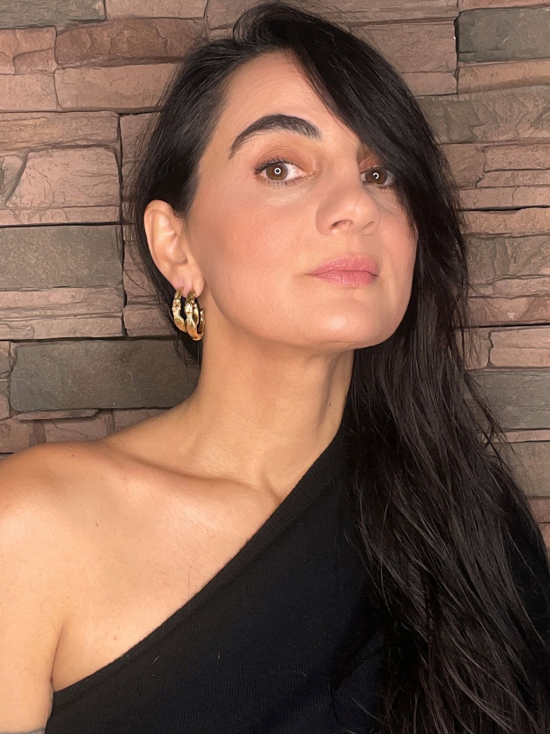 Woman with long dark hair wearing gold sculptural goddess hoop earrings against a stone wall background