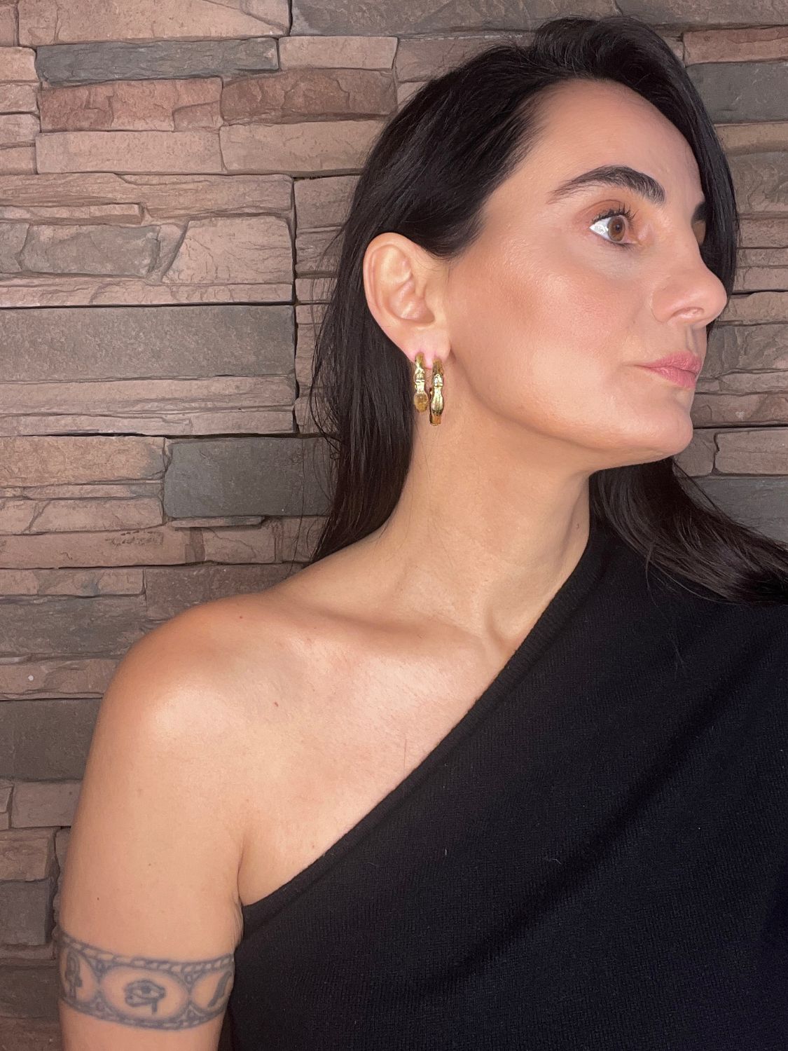 Woman wearing a black top with sculptural gold hoops against a stone wall background