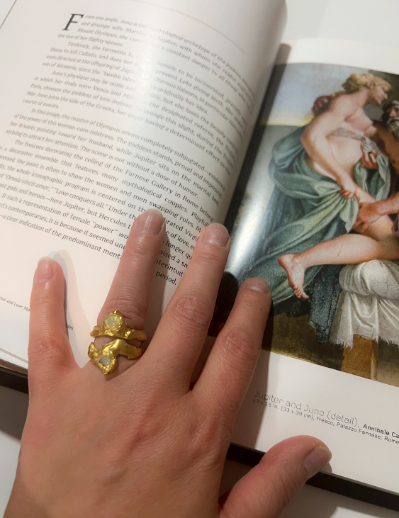 A person's hand displaying a gold ring with a faceted fire opal on the ring finger, with a book in the background featuring a classical painting.