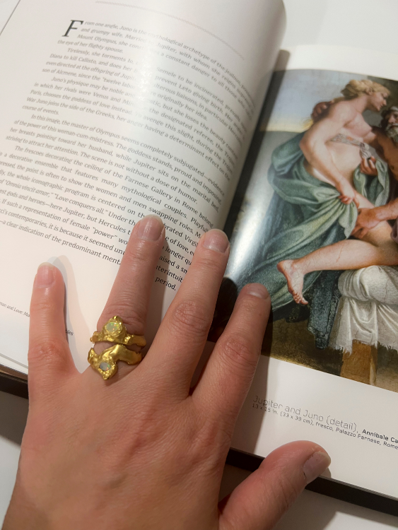 A person's hand displaying a gold ring with a faceted fire opal on the ring finger, with a book in the background featuring a classical painting.